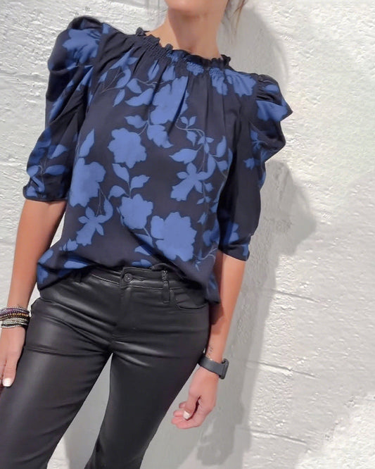 Person wearing a blue floral blouse and black pants against a light gray wall.