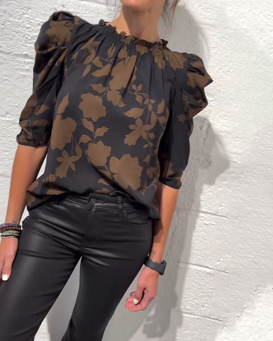 Person wearing a black floral blouse and black pants against a white wall